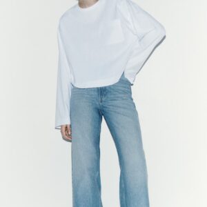 Agrandir l'image: Un haut blanc à manches longues présente un col rond et des poignets froncés, avec le devant partiellement rentré. Il est porté avec un jean bleu clair à taille haute et à jambes larges, mettant en valeur un ourlet effiloché et des poches avant visibles, légèrement orienté vers la droite.