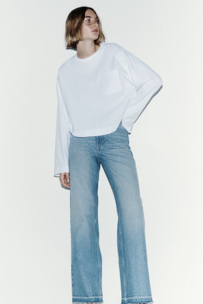 Agrandir l'image: Un haut blanc à manches longues présente un col rond et des poignets froncés, avec le devant partiellement rentré. Il est porté avec un jean bleu clair à taille haute et à jambes larges, mettant en valeur un ourlet effiloché et des poches avant visibles, légèrement orienté vers la droite.
