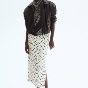 Une jupe midi ajustée en jersey, arborant un motif à pois noirs sur un fond clair, est portée par le mannequin. La jupe présente une taille haute, tombe délicatement sur les côtés et se termine par un ourlet droit avec une fente haute sur un côté.