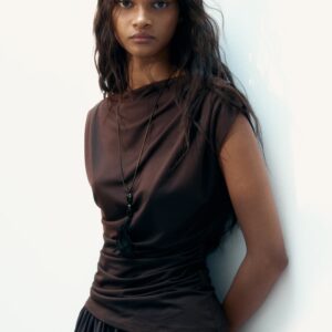 Le modèle porte un haut en jersey marron foncé asymétrique, doté d'un col bateau, de manches courtes avec plis aux épaules, et de fronces sur les côtés à la taille créant un effet drapé. Un long collier noir avec un pendentif à pompon est superposé sur le haut.